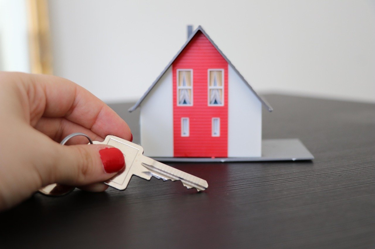 Person holding house keys beside a small red-and-white model home, representing purchase of a short lease property.